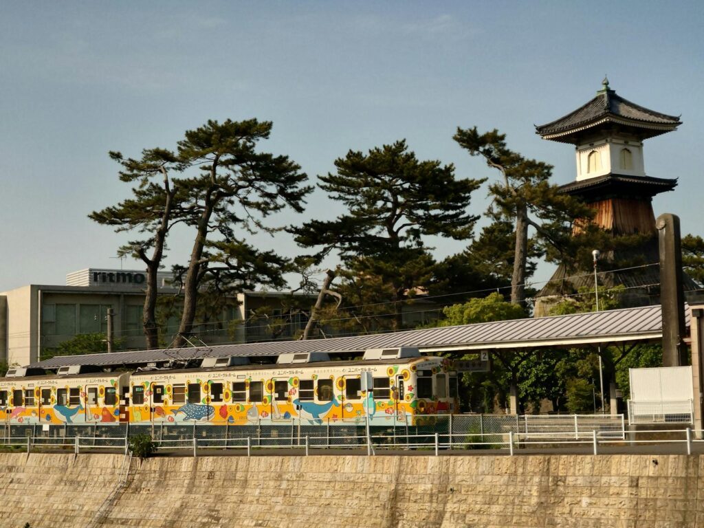 A vibrant train travels through a scenic area beside traditional Japanese architecture and trees.